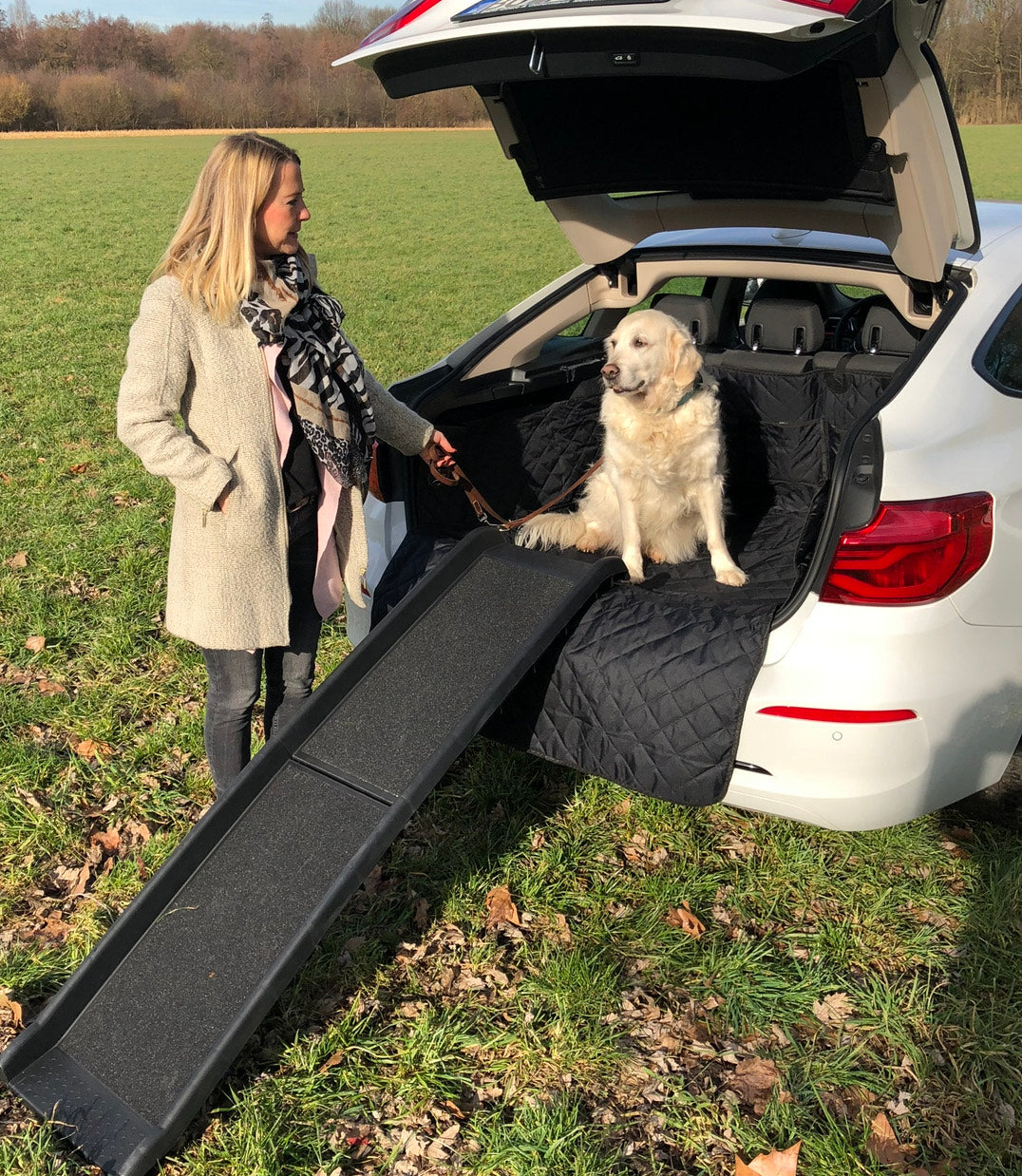 Eine Frau steht neben einem Auto mit Hunderampe wo ein weißer Hund im Kofferraum sitzt